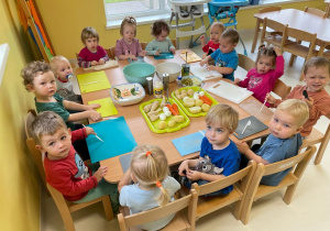 Zdjęcie dzieci z grupy Żabki siedzących przy stoliku przed robieniem sałatki warzywnej.