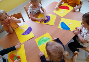 Grupa dzieci siedzących przy stole przykleja kolorowe parasole na kartki papieru.