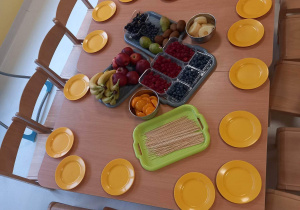 Zdjęcie stołu z przygotowanymi owocami, patyczkami szaszłykowimi i talerzykami dla dzieci.