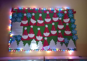 Ozdobiona kolorowymi świecącymi lampkami, tablica z wykonanymi Świętymi Mikołajami przez dzieci z grupy Pszczółki.