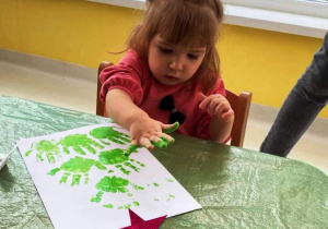 Maja podczas odciskania na kartce pomalowanej rączki na zielono.
