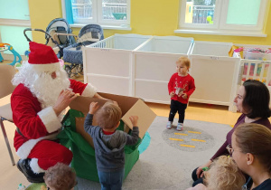 Zdjęcie Świętego Mikołaja otwierającego razem z dziećmi z grupy Motylki wielkie zielone pudło z prezentami.