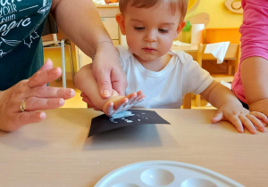 Opiekunka z grupy Motylki pomaga Jasiowi odcisnąć dłoń pomalowaną białą farbą na czarnej kartce papieru.