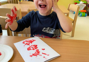 Zadowolony Maciek podczas zajęć, pozuje do zdjęcia z pomalowaną ręką na czerwono.