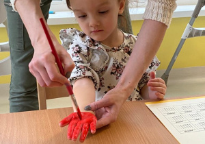 Opiekunka maluje dłoń dziewczynki za pomocą pędzelka i czerwonej farby.