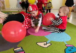 Zdjęcie maluchów siedzących na dywanie i bawiących się czerwonymi balonami.