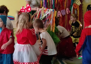 Fotografia dzieci przechodzących pod ozdobioną kolorowymi bibułami tyczką.