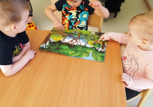 Troje dzieci siedzących przy stoliku ogląda książeczkę z dinozaurami.