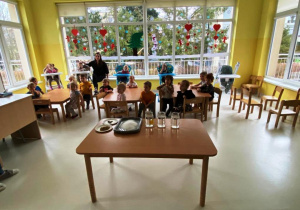 Fotografia przedstawiająca wszystkie dzieci oraz stół z akcesoriami do zabawy badawczej.