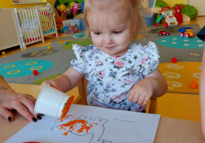Wiktoria dekorująca swojego dinozaura kubeczkiem z pomarańczową farbą.