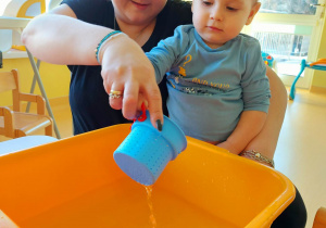 Opiekunka pomaga Szymonowi wylać wodę z niebieskiego kubeczka do żółtej miski.