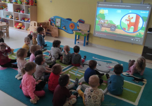 Dzieci z grupy Pszczółki siedzą na dywanie i oglądają na tablicy interaktywnej bajkę edukacyjną pt. ,,Zwierzęta leśne''.