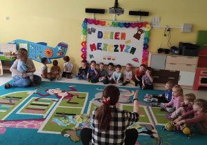 Zdjęcie opiekunki, opowiadającej siedzącym dziewczynkom i chłopcom na dywanie o Dniu Mężczyzn.