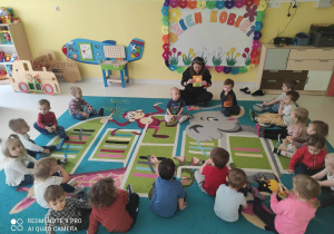 Zdjęcie dzieci siedzących w kółku na dywanie razem z opiekunką, która opowiada o Dniu Kobiet i pokazuje wykonane własnoręcznie laurki przez chłopców.