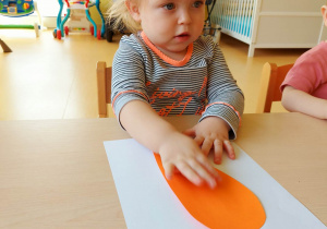 Laura przykleja na białą kartkę A4, wycięty z pomarańczowej kartki szablon korzenia marchewki.