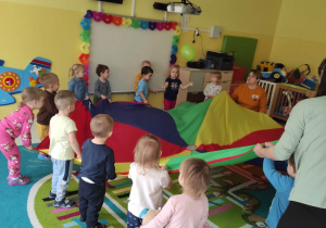 Zdjęcie dzieci i opiekunek z grupy Pszczółki podczas zajęć na dywanie z chustą animacyjną i zielonym balonikiem.