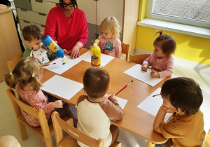 Dzieci siedzące przy stole z pomocą opiekunki mieszają na kartce dwa kolory farb.