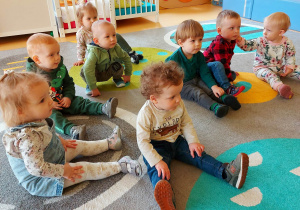 Dzieci z grupy Motylki siedzą na dywanie i słuchają opiekunki opowiadającej o zwiastunach wiosny i pokazującej wiosenne obrazki w książce.