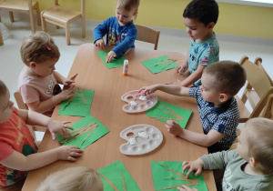 Dzieci siedzące przy stoliczku przyklejają na zielone kartki papieru łodyżki bazi wierzbowych.