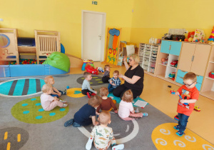 Zdjęcie dzieci i opiekunki z grupy Motylki siedzących na dywanie podczas pogadanki pt. ,,Co to jest rytm?''.