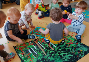Zdjęcie dzieci siedzących na podłodze podczas ozdabiania ich wspólnej pracy plastycznej kolorowymi kwadracikami z bibuły.