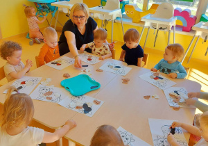 Zdjęcie siedzącej przy stoliku obok dzieci opiekunki, trzymającej w dłoni czarną plamkę wyciętą z bloku rysunkowego.