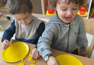 Zdjęcie Wiktora i Brunona podczas przyklejania żółtej bibuły na papierowe żółte talerzyki.