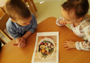 Dwoje dzieci przy stole wpatruje się w ilustrację koszyczka wielkanocnego.