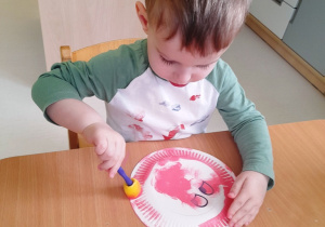 Aleksander stempluje gąbką umoczoną w różowej farbie papierowy talerzyk.