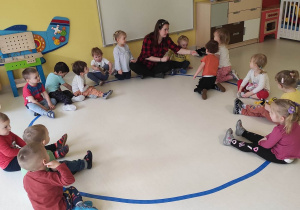 Zdjęcie opiekunki i dzieci siedzących na podłodze na niebieskim kręgu zrobionym z niebieskiej taśmy podczas zajęć pt. ,,Memory''.