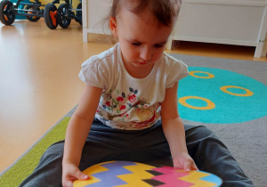 Zdjęcie siedzącej na dywanie Barbary próbującej złączyć ze sobą dwuelementowe puzzle w kształcie kolorowej pisanki.