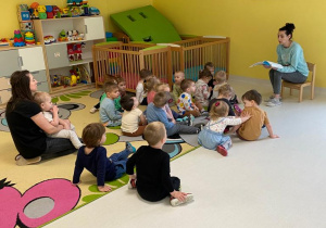 Dzieci siedząc na dywanie słuchają wiersza czytanego przez opiekunkę.