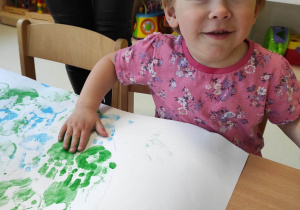 Alicja uśmiecha się do zdjęcia podczas odciskania swojej dłoni namoczonej w zielonej farbie.