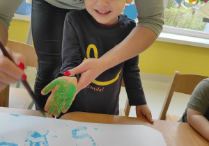 Hubert uśmiecha się do zdjęcia podczas malowania przez opiekunkę jego dłoni zieloną farbą.