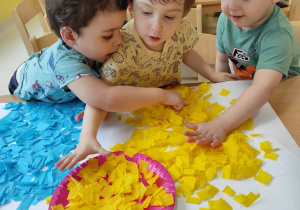 Artur, Wiktor i Oliwier wspólnymi siłami ozdabiają kubeł kwadracikami wyciętymi z żółtej bibuły.