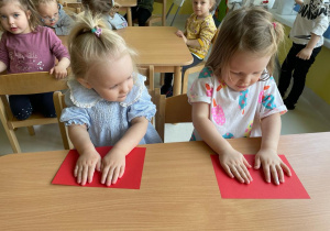 Dwie dziewczynki odciskają swoje dłonie pomalowane białą farbą na czerwonej kartce.