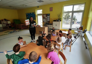 Dzieci siedzące przy stole słuchają opiekunki, która opowiada o godle Polski.