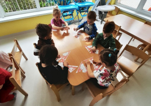 Grupa dzieci siedząc przy stole układa puzzle przedstawiające białego orła w koronie na czerwonym tle.