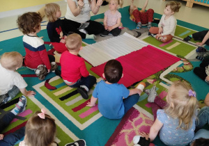 Opiekunka pokazuje siedzącym na dywanie dzieciom serduszka wycięte z białego i czerwonego papieru.