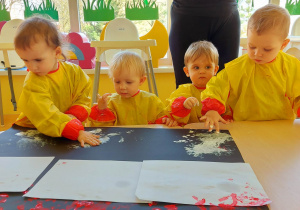 Zdjęcie dzieci odciskających swoje rączki pomalowanych białą farbą na czarnym brystolu.