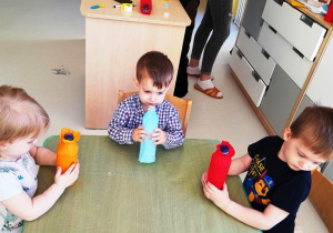 Troje dzieci siedzących przy stole z obklejonymi bibułą butelkami.