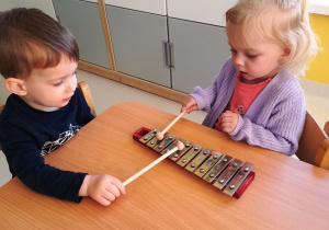 Dwoje dzieci siedzących przy stole stukają pałeczkami w dzwonki chromatyczne.