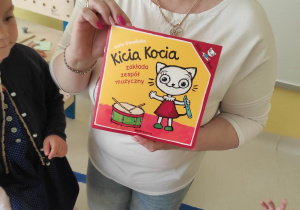 Zdjęcie opiekunki trzymającej w dłoniach książeczkę pt.,,Kicia Kocia zakłada zespół muzyczny''.