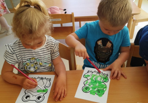 Dwoje dzieci siedzących przy stoliku malują żabki zieloną farbą.