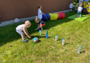 Dzieci podczas zabawy na dworze zbijają piłką kręgle zrobione z plasttikowych butelek.