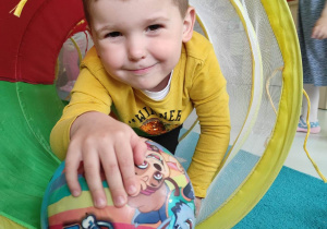 Uśmiechnięty Oliwier wychodzi z tunelu trzymając w dłoni kolorową piłkę.