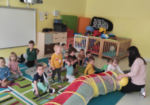 Zdjęcie opiekunki i dzieci siedzących na dywanie przygotowujących się do zajęć z kolorowym tunelem.