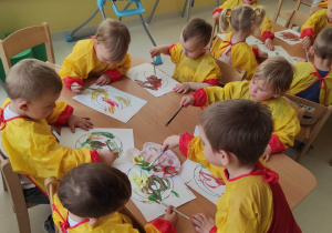 Dzieci siedzące przy stoliczku malują kolorowymi farbami swoje piłeczki.