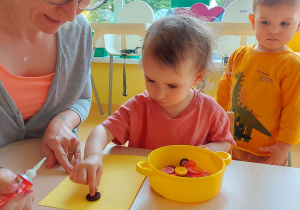 Barbara starannie przykleja na swoją żółtą kartkę guzik w kolorze ciemno-fioletowym.
