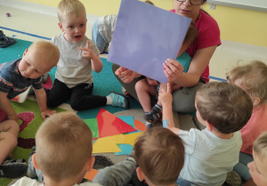 Opiekunka z grupy Pszczółki pokazuje dzieciom fioletowy kwadrat podczas zajęć pt. ,,Co można ułożyć z figur?''.